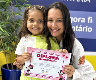Menina com diploma rosa ao lado de uma profissional na clínica.