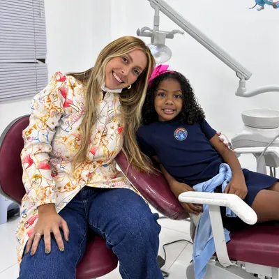 Profissional de jaleco estampado ao lado de uma menina na cadeira odontológica.