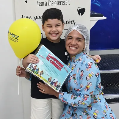 Menino com certificado e balão amarelo ao lado de uma profissional.
