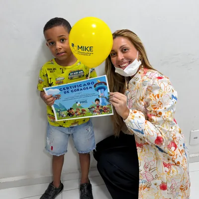 Menino com certificado e balão ao lado de uma profissional de jaleco estampado.