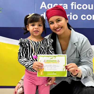 Menina com certificado ao lado de uma profissional com touca rosa.